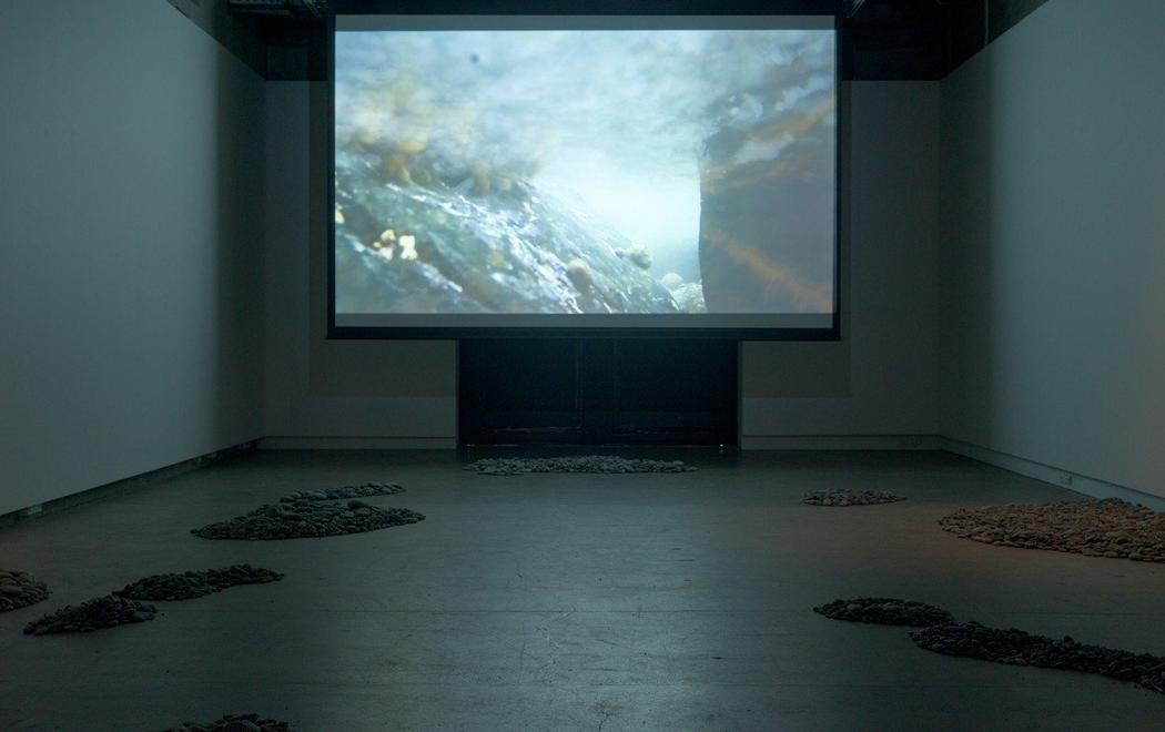 A screen plays an underwater image in a darkened room. There are shaped piles of stones in the foreground.