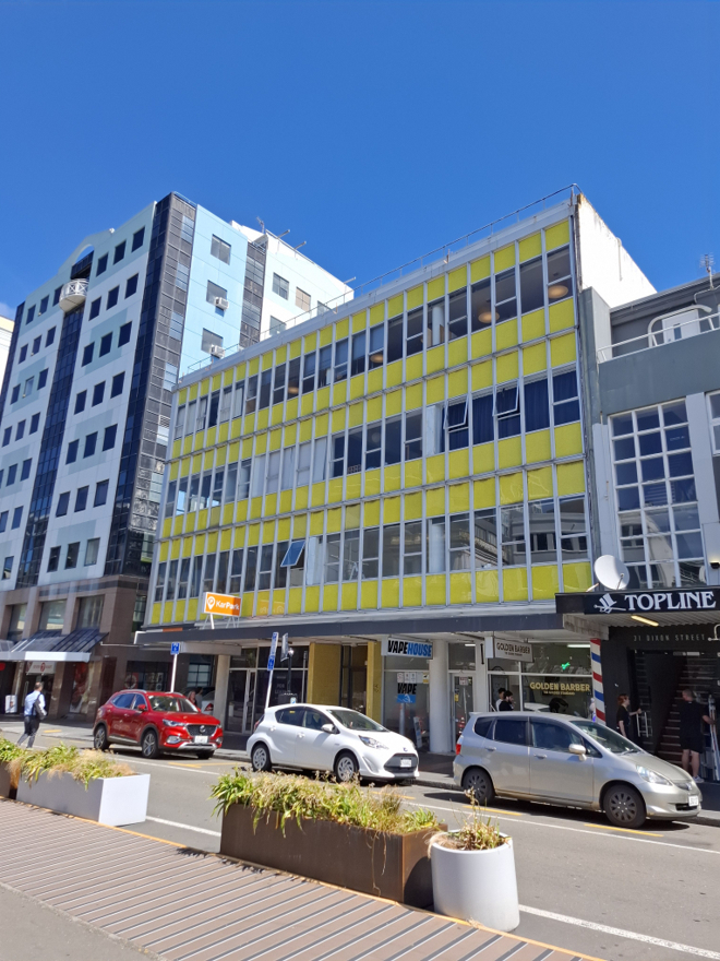 A 1960s modernist office building with yellow panelling and large windows. Cars are parked in the street below.