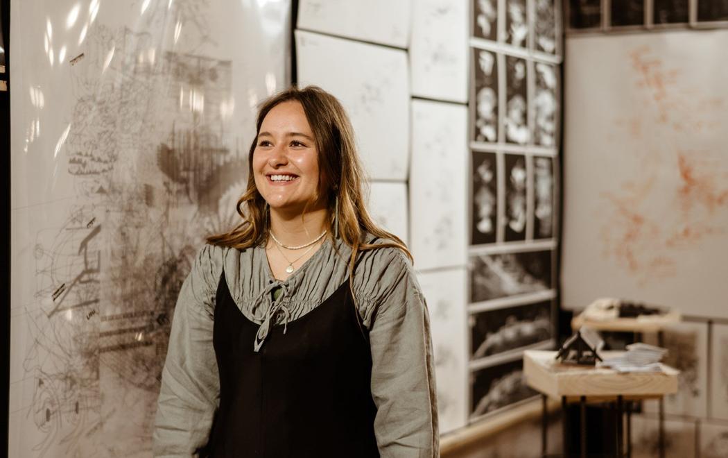 Portrait of a smiling young woman; an exhibition or active studio can be seen in the background.