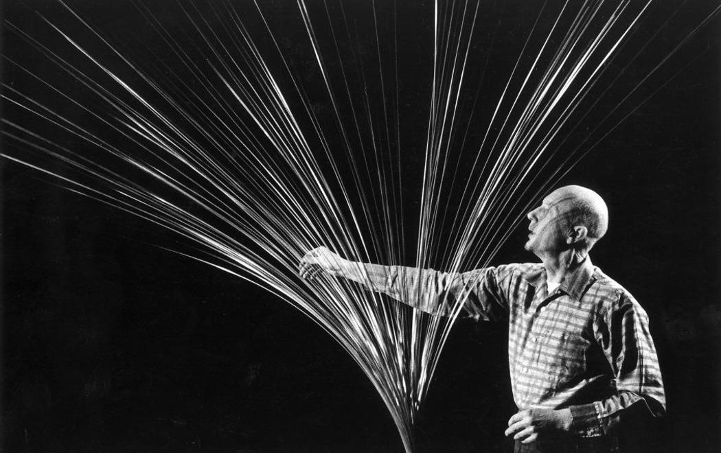 Artist Len Lye reaches towards a sculpture made of metal rods fanning out from the base. Lye's gaze is fixed as if to gauge the movement of the metal rods, and his had raches as if to steady one of the rods.
