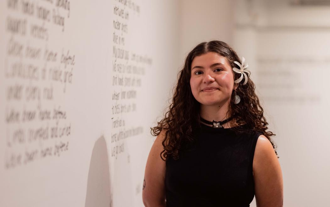 A young woman in semi-formal attire stands besdie a gallery wall. She is smiling and has a flower in her hair. On the wall is blurry text.