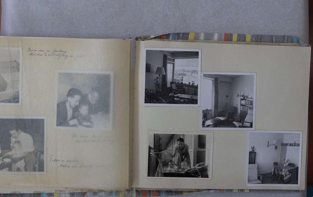A family photo album is open on a table. The photos are black-and-white and depict domestic interiors alongside scenes of family life.