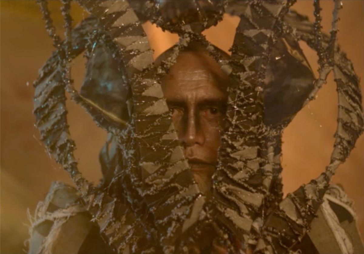 The face of a male figure shows a stern expression. Their head is enveloped by a series of green wavy tendrils, which point upright.