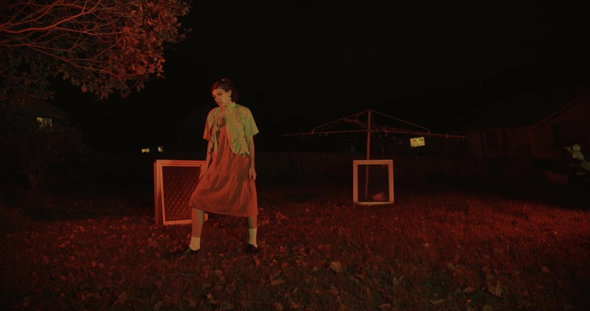 In a backyard a person in a dress stands in front of two windows which are propped up on the lawn. The second window rests against a clothes line. The scene is lit by a red glow.